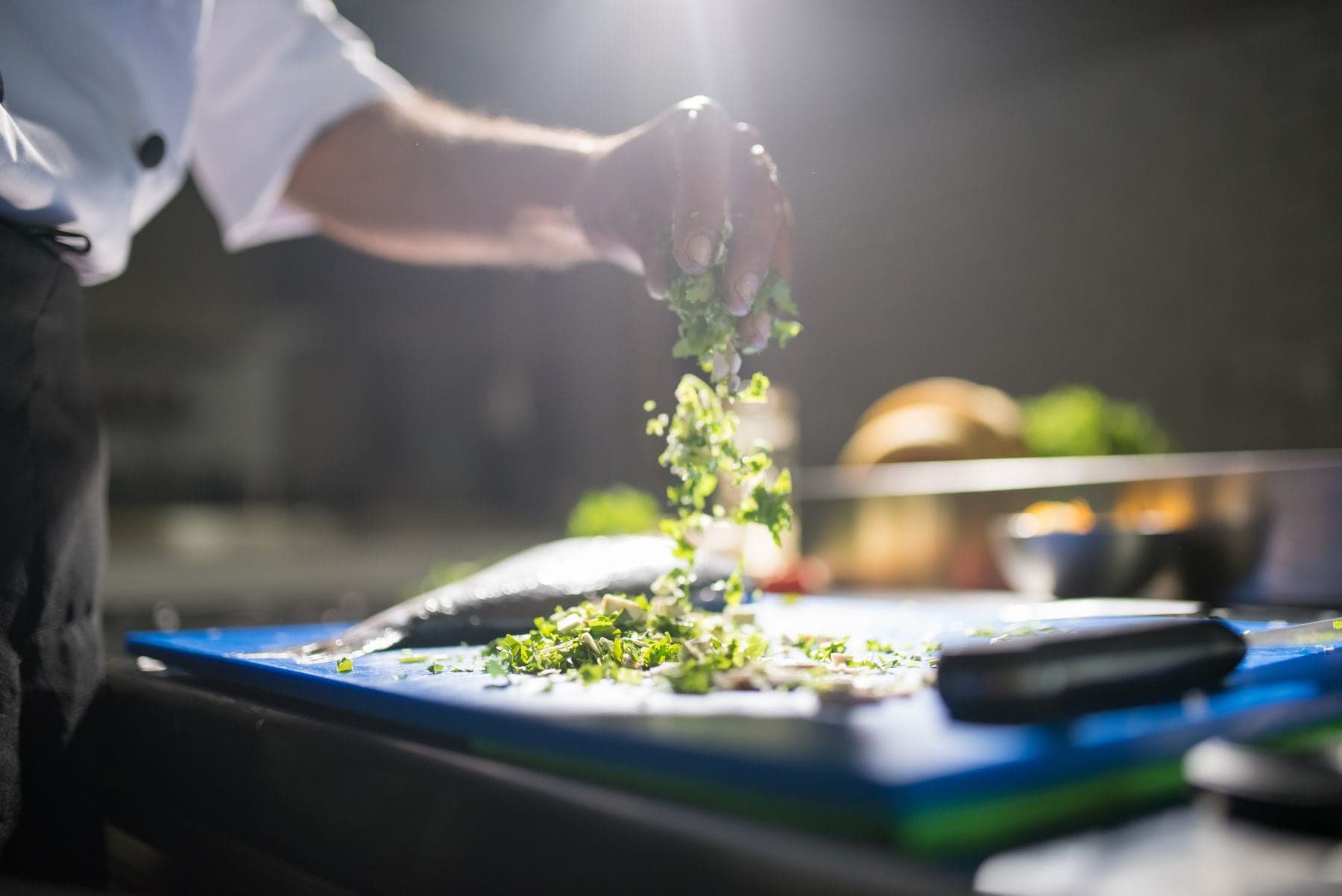 A cook dropping chopped herbs onto a board in a dark kitchen