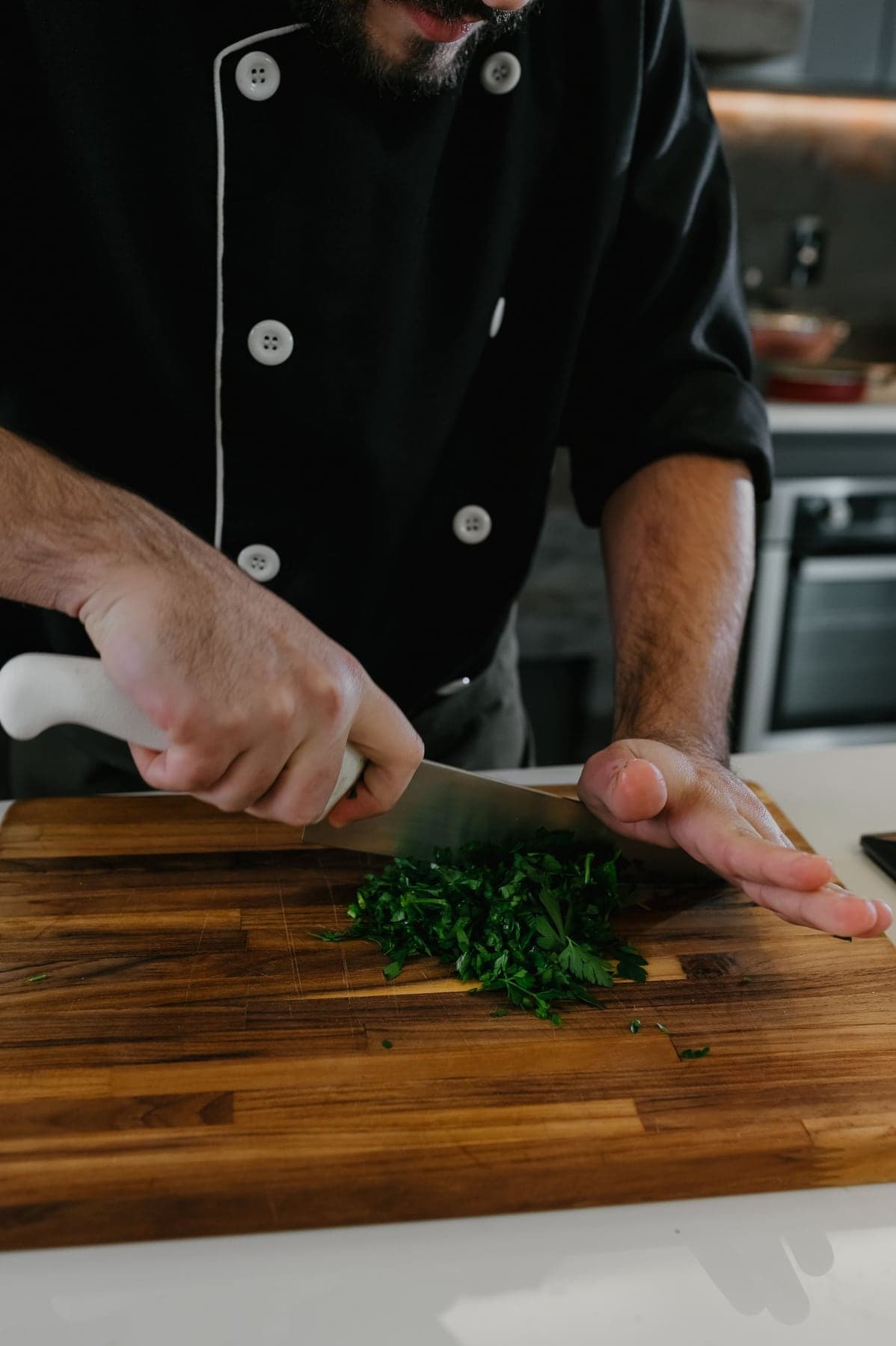 A chef in a black jacket chopping fresh herbs on a wooden board