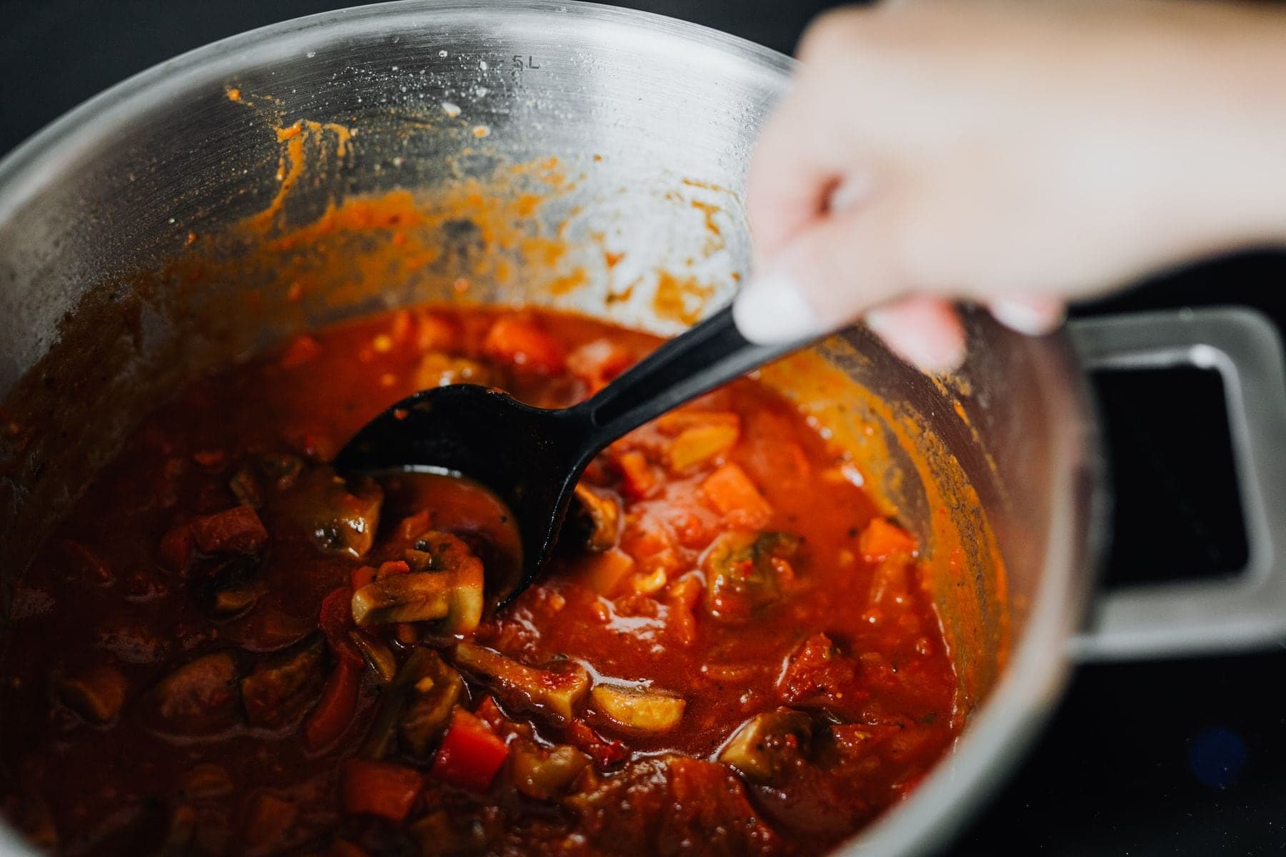 A hand stirring a pot of tomato stew with a spoon