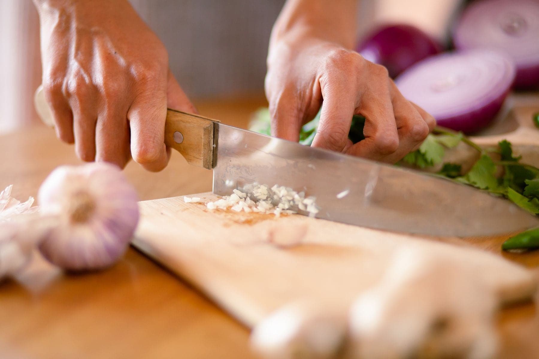 Hands mincing garlic on a wooden board beside red onions